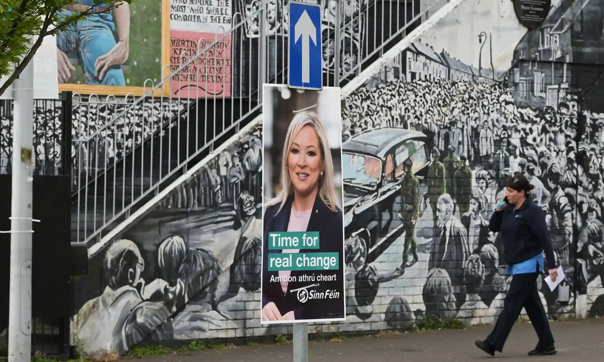 ‘Ώρα για πραγματική αλλαγή’ > αφίσα του Sinn Féin, με την αντιπρόεδρο Michelle O’Neill μπροστά από ρεπουμπλικανική τοιχογραφία στο δυτικό Belfast / Charles McQuillan, Getty Images, theguardian.com